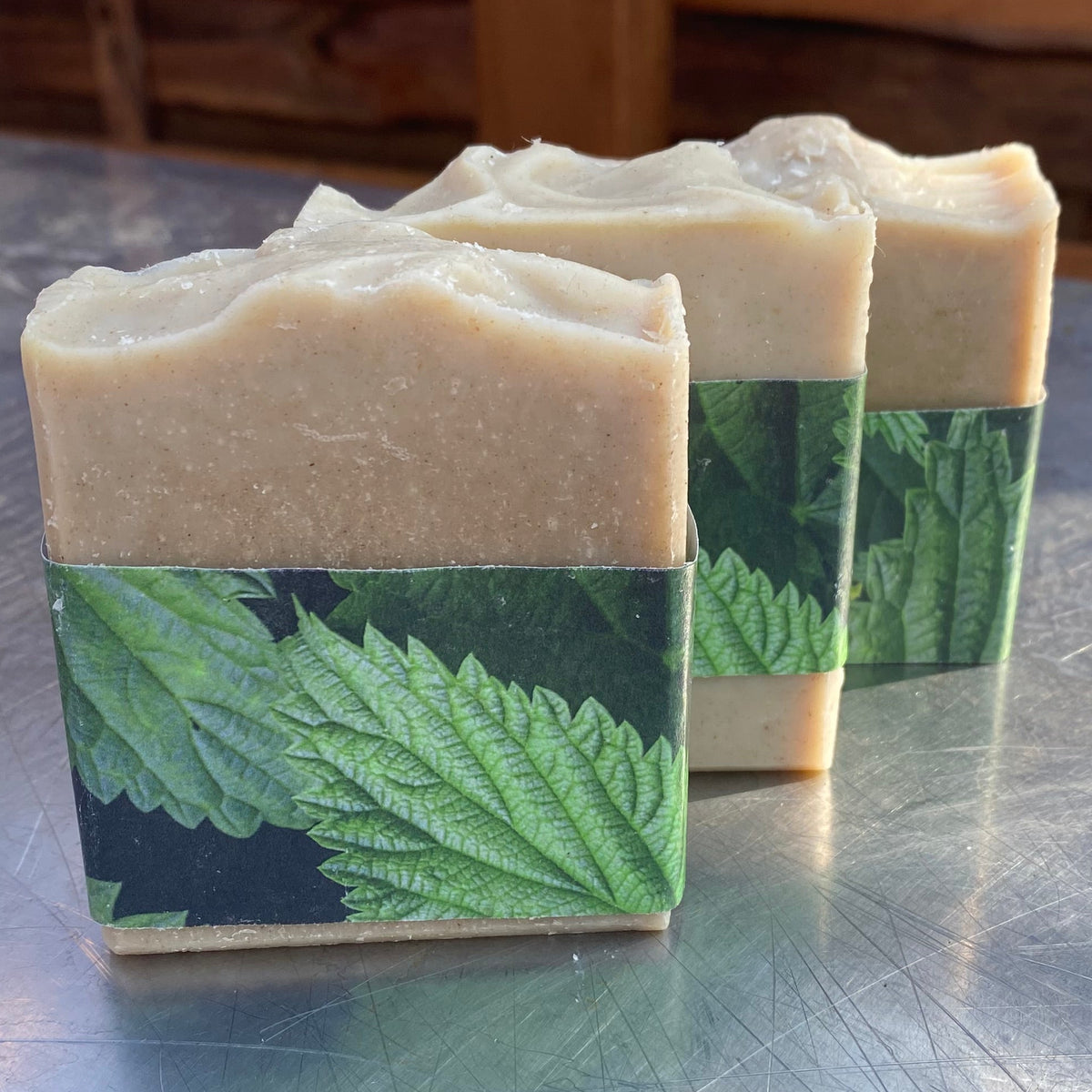 Three handmade soap bars with a light beige top and a green leaf patterned paper sleeve, standing on a scratched silver metal surface.