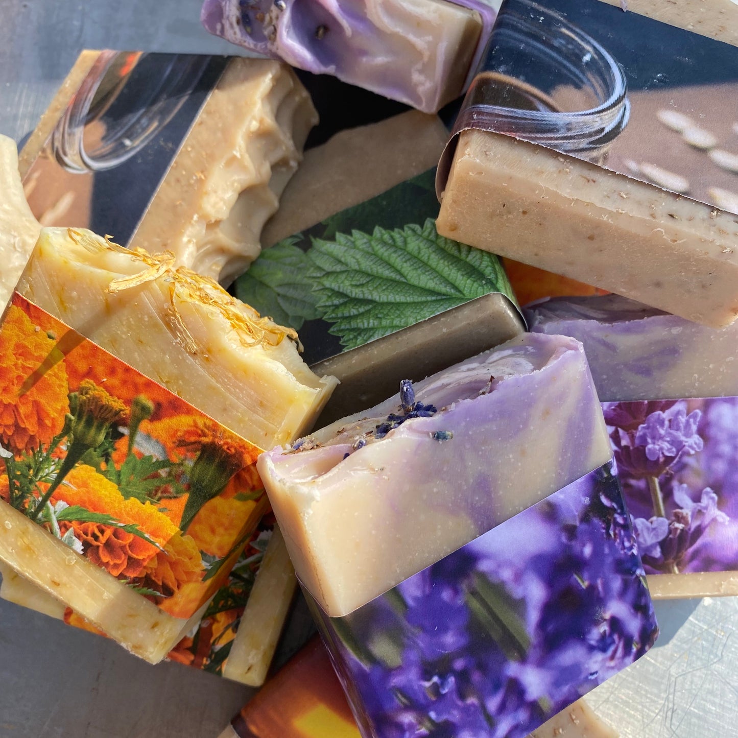 A pile of artisanal handmade soaps with floral and botanical designs, including purple lavender, orange marigold, and green leaf patterns, arranged on a metallic surface.