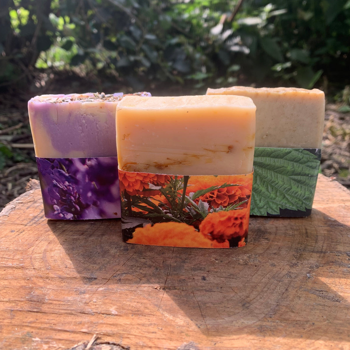 Three rectangular handmade soaps with colorful botanical and floral printed paper sleeves, displayed on a rustic wooden log with a blurred green outdoor background.