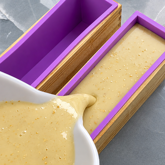 A close-up of a white bowl pouring pale yellow soap batter into a purple silicone mold with a bamboo frame, with another empty purple mold beside it on a gray stone surface.