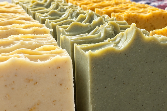 Multiple rectangular bars of handmade soap in pale yellow, sage green, and orange-yellow, arranged in rows on a wooden surface against a white brick wall background.