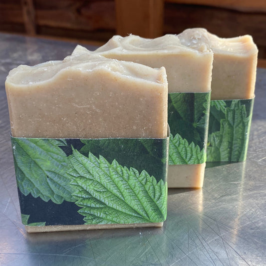 Three handmade soap bars with a light beige top and a green leaf patterned paper sleeve, standing on a scratched silver metal surface.