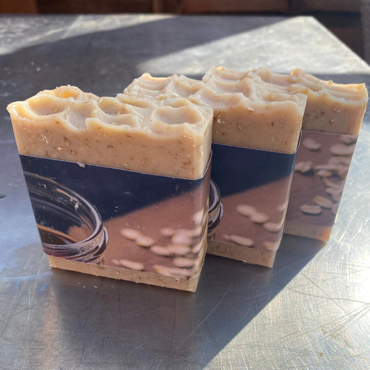 Three rectangular bars of handmade soap with a dark navy blue label featuring a picture of a glass jar and beans, standing on a scratched metal surface.
