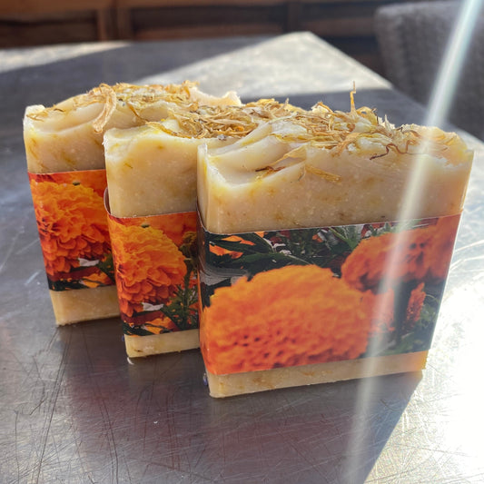 Three handmade rectangular bars of soap with orange marigold floral labels and dried botanicals on top, arranged on a scratched metal table.