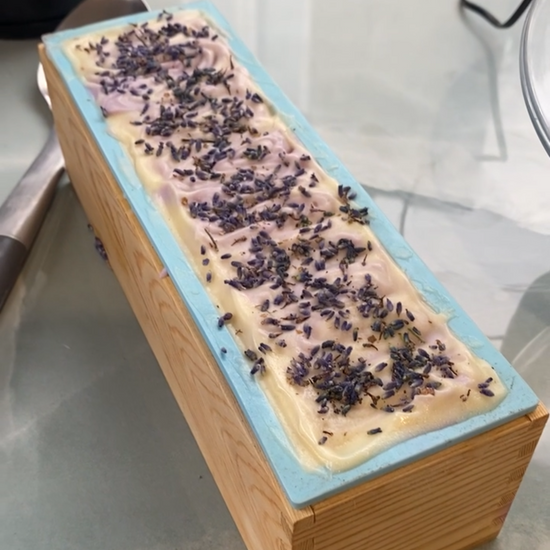 A rectangular bar of soap in a wooden mold, featuring a pale pink base with dark purple dried flower petals on top and a light blue edge, photographed on a glass tabletop.