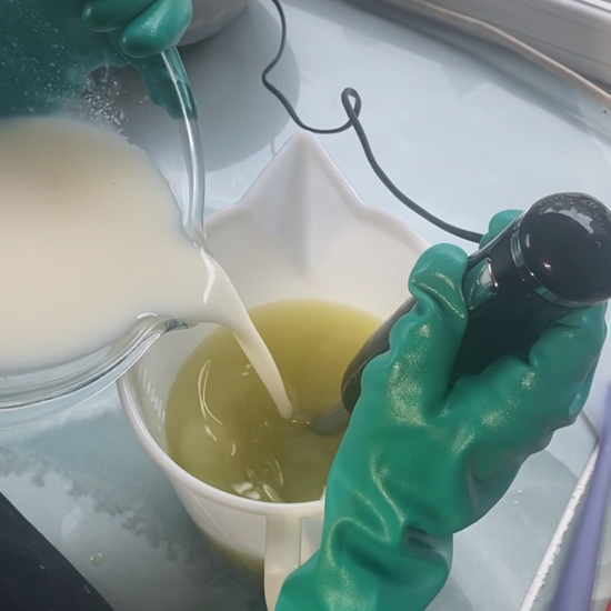 A person wearing green rubber gloves pours a white liquid into a bowl containing a pale yellow soap mixture, with a metal bowl of wax chunks nearby on a reflective tabletop.