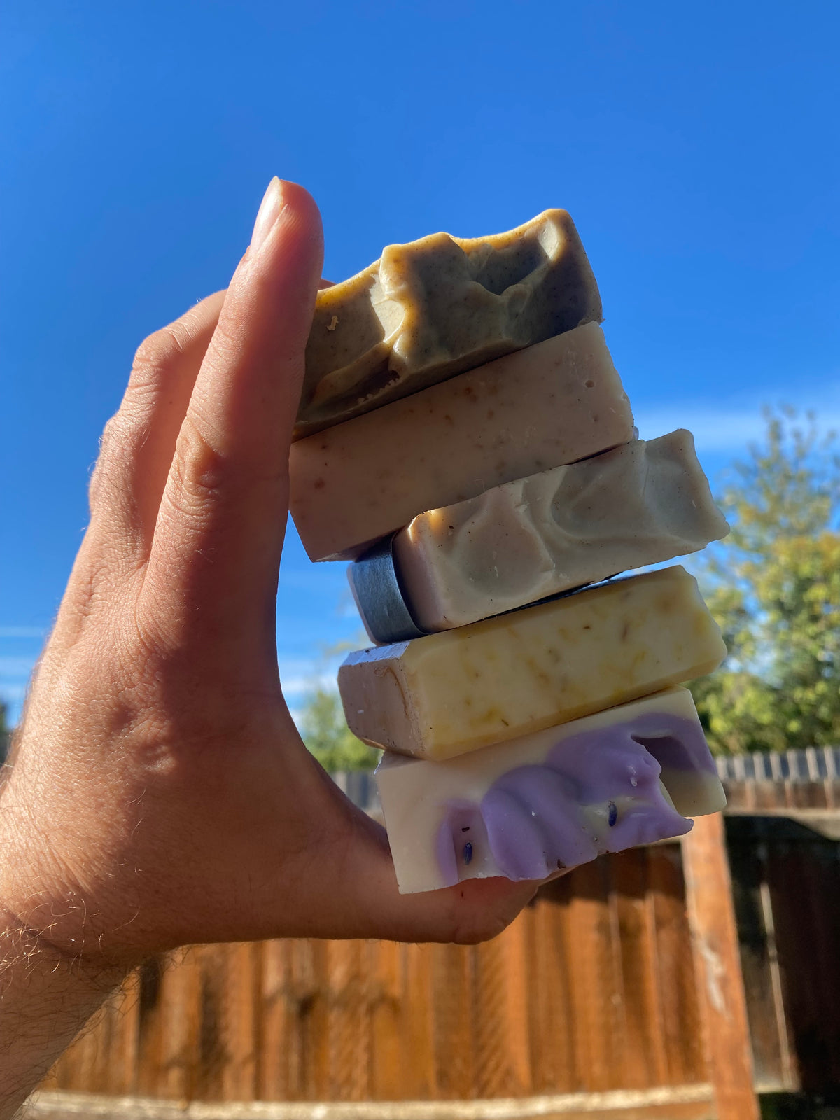 A hand holds five handmade soap bars in different colors and patterns against a bright blue sky and wooden fence background.