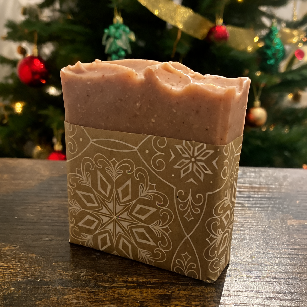 A rectangular bar of handmade soap in a brown paper sleeve with a white snowflake pattern, sitting on a dark wooden table in front of a decorated Christmas tree.