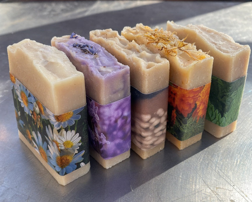 Five handmade artisanal soaps with colorful floral and nature-themed paper bands, arranged on a scratched metallic surface with a rustic wooden background.