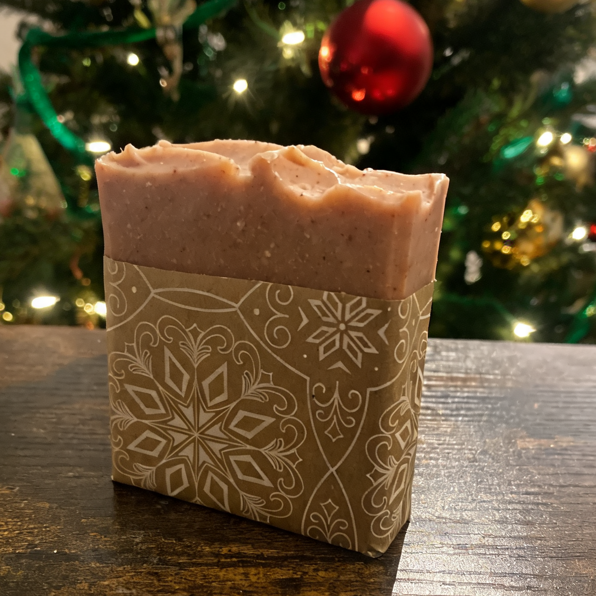 A rectangular bar of pinkish-beige soap wrapped in a brown paper sleeve with a white snowflake pattern, sitting on a dark wooden table with a blurred Christmas tree in the background.